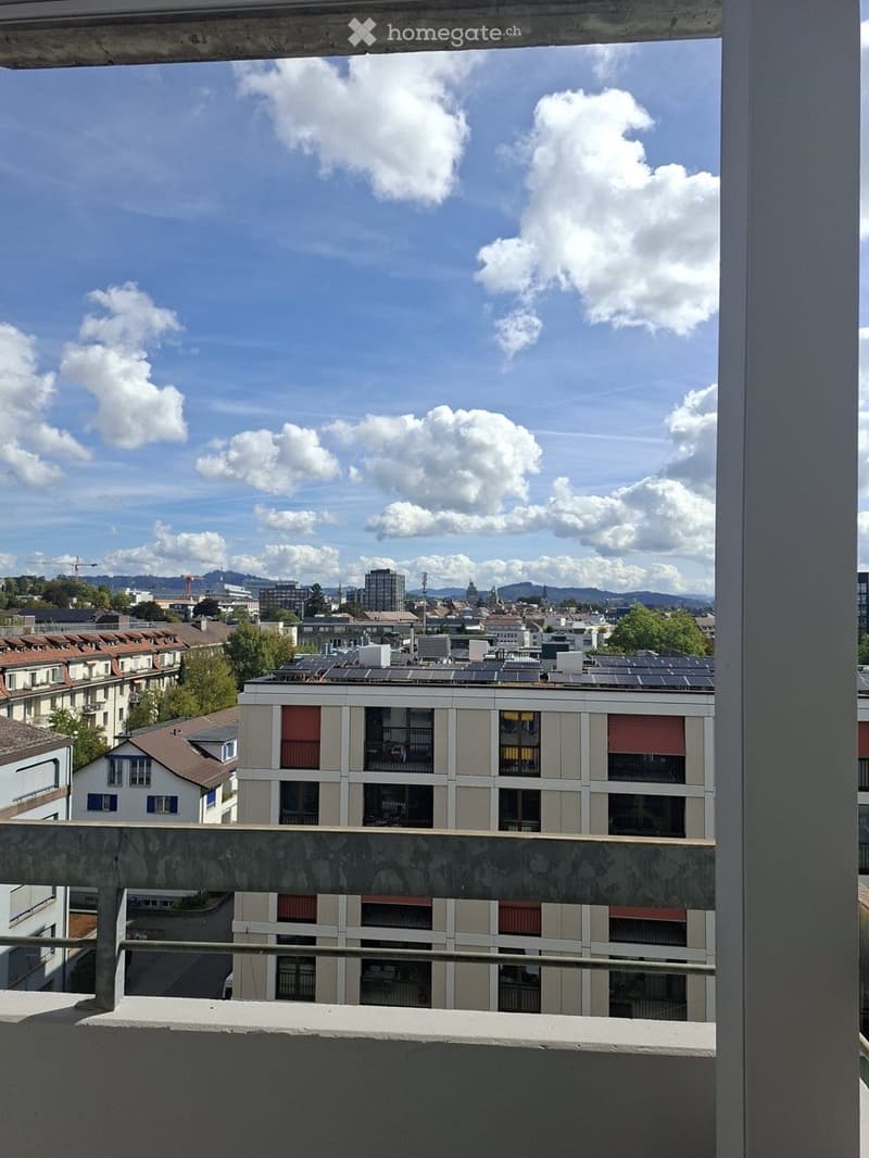 Balcony overlooking a cityscape with buildings, solar panels on rooftops, and a partly cloudy sky.