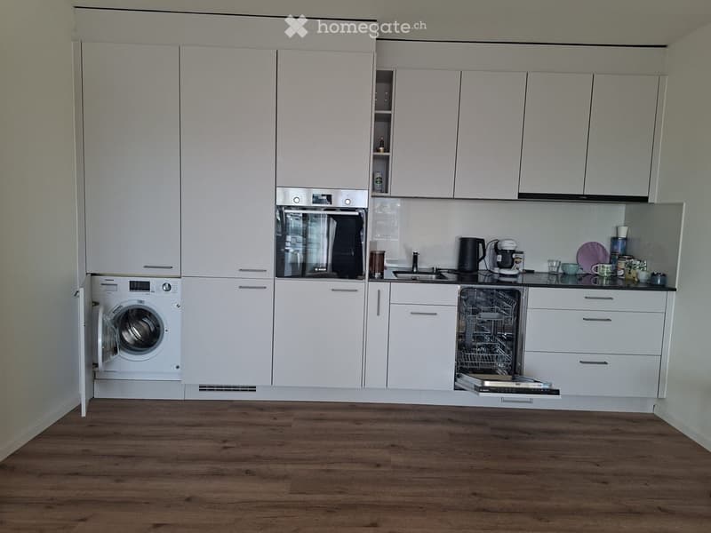 Modern kitchen with white cabinets, wooden floor, and integrated appliances including a washing machine, dishwasher, and oven. Countertop features a coffee maker, cups, and a bowl.