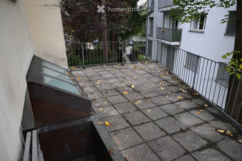 Balcony with concrete tiles, metal railing, scattered leaves, and a small roofed area on the left.