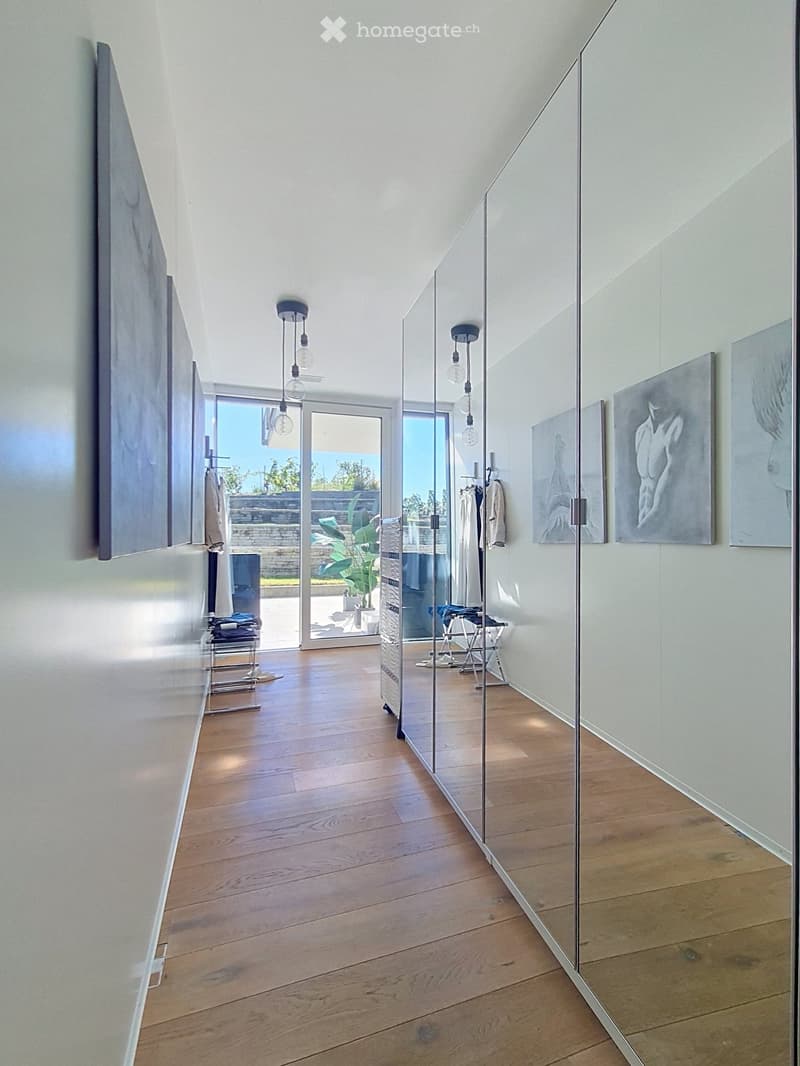 Modern hallway with wooden flooring, glass walls, and pendant lights. Features artwork on the walls and a view of an outdoor area through large glass doors.