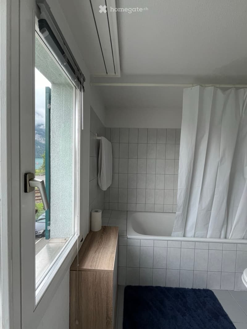 Compact bathroom with a bathtub, toilet, shower curtain, and a small wooden shelf holding a towel and toilet paper. A window offers a view of the outside.