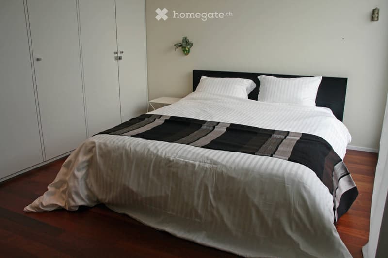 Modern bedroom with a large bed, nightstand, and wall-mounted lamp. The bed features a black headboard and a patterned duvet cover.
