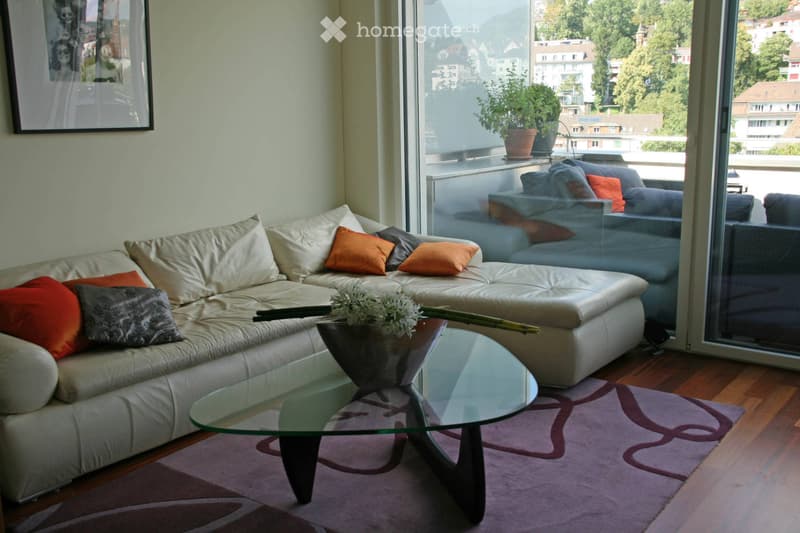 Modern living room with a white leather sofa, glass coffee table, and a view of the city and mountains through large windows.