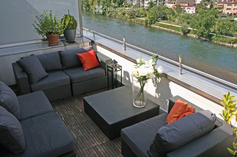Balcony with a view of a river, featuring a grey wicker sofa, a small table with a vase of white flowers, and potted plants.