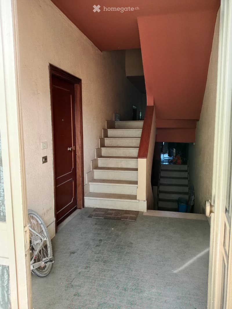 Hallway with tiled floor, staircase leading upwards, wooden door on left, bicycle leaning against wall, small rug near stairs.