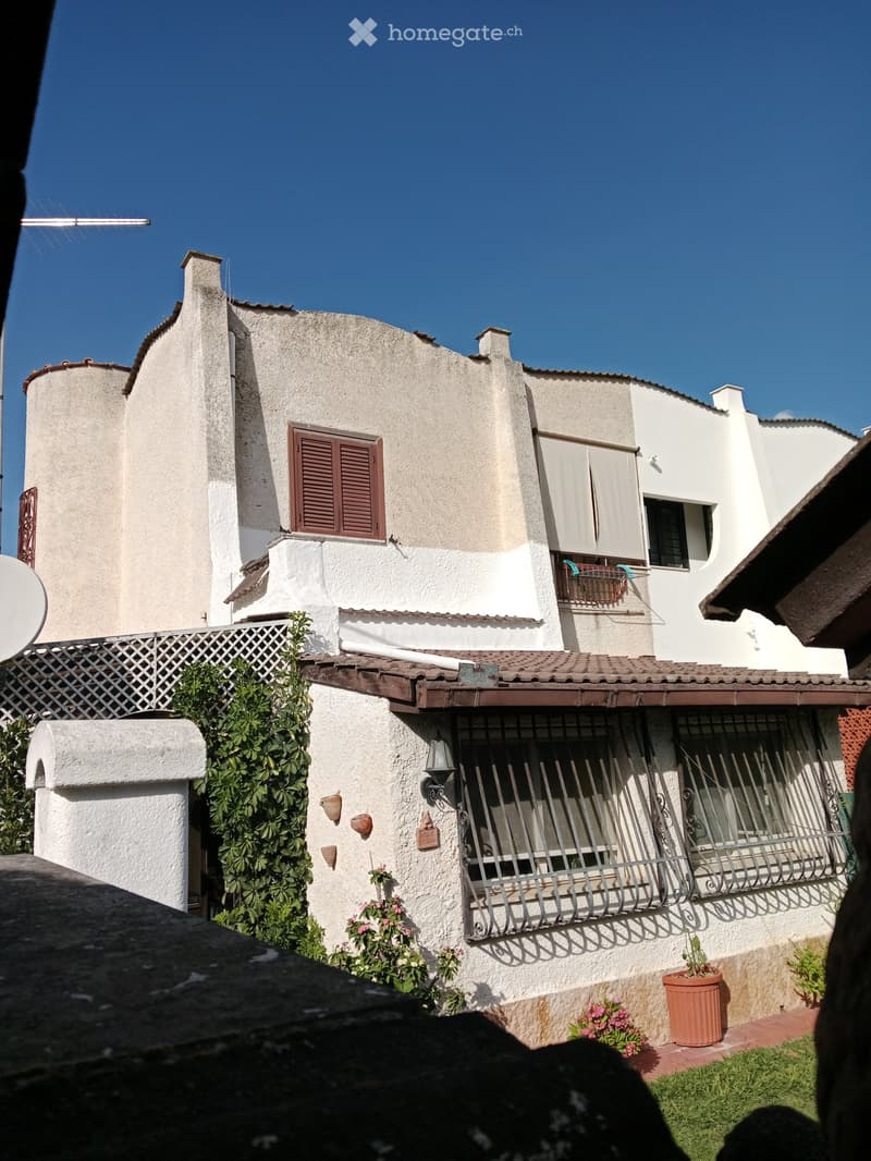 Two-story house with beige and white exterior, brown shutters, metal balcony railings, and small garden area with potted plants.