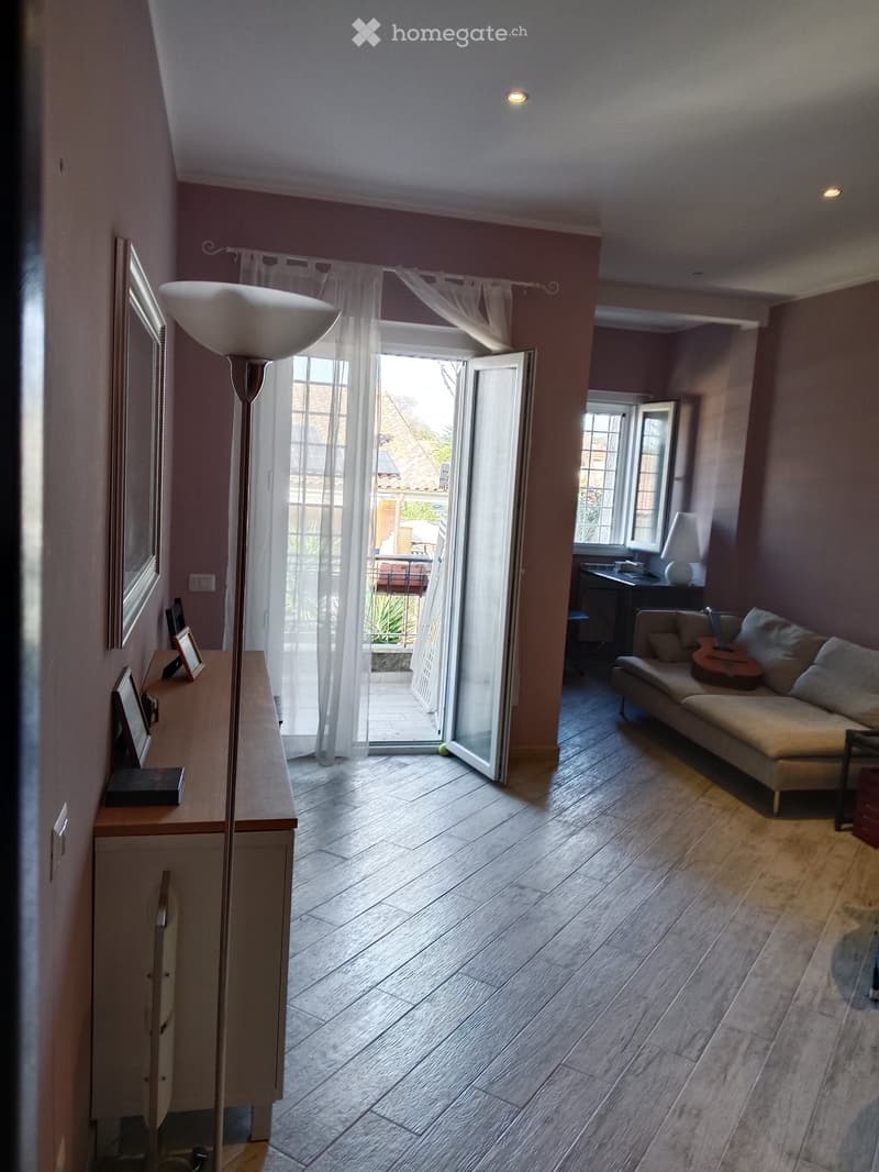 Living room with wooden flooring, large window with sheer curtains, open door leading to a balcony, sofa with guitar, side table with picture frames, and a floor lamp.