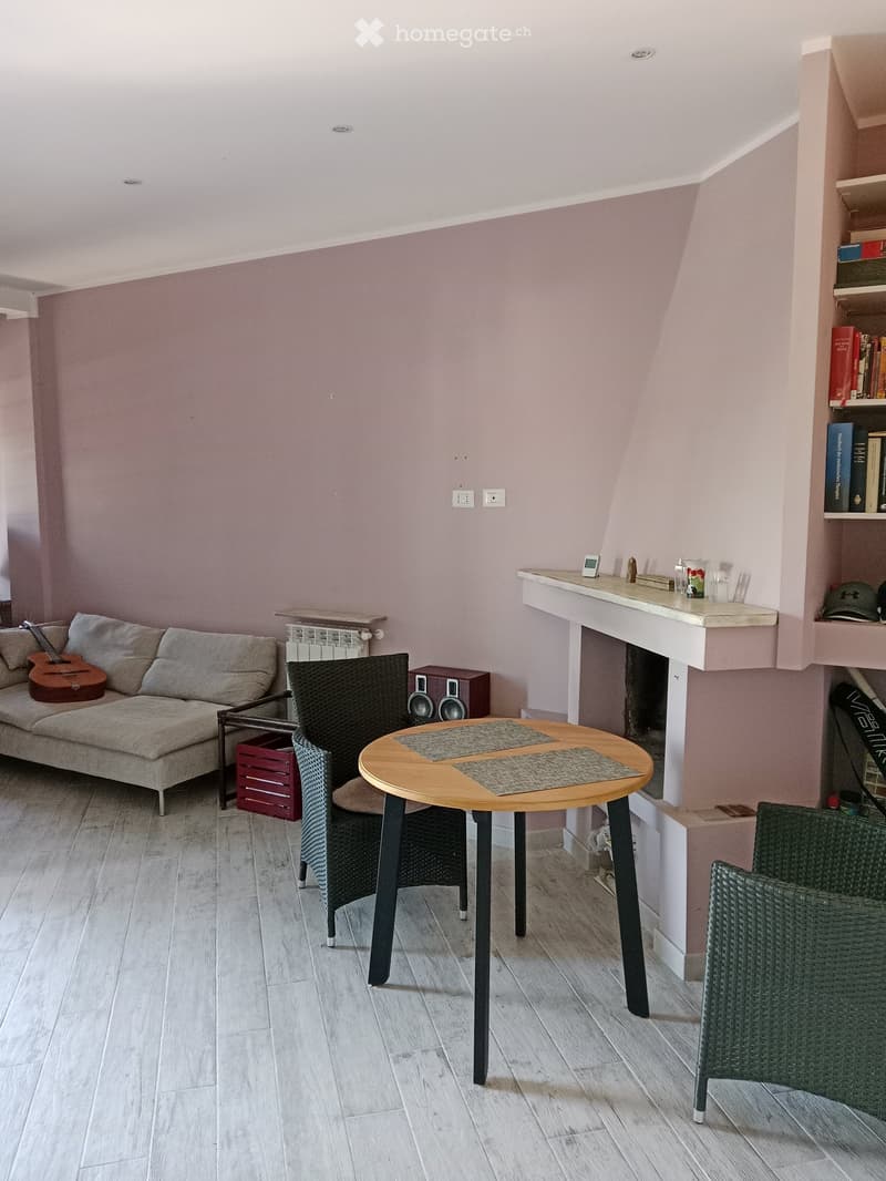 A cozy living room with a grey sofa, a round wooden table, wicker chairs, a guitar, and a speaker system. The room features light pink walls, wooden flooring, and built-in shelving with books and decorative items.