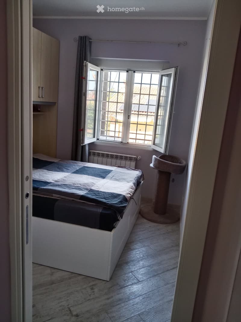 Bright bedroom with a white bed, a cat tree, a radiator, and a cabinet. Large windows with open shutters let in natural light.