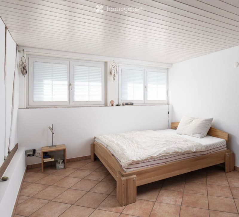 Bright bedroom with wooden bed, nightstand, and dreamcatchers on the wall. The room features large windows with blinds and a tiled floor.