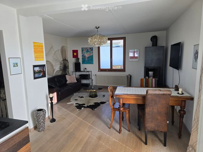Modern living room with wooden flooring, a black leather sofa, a wooden dining table with chairs, a fireplace, and decorative artwork on the walls.