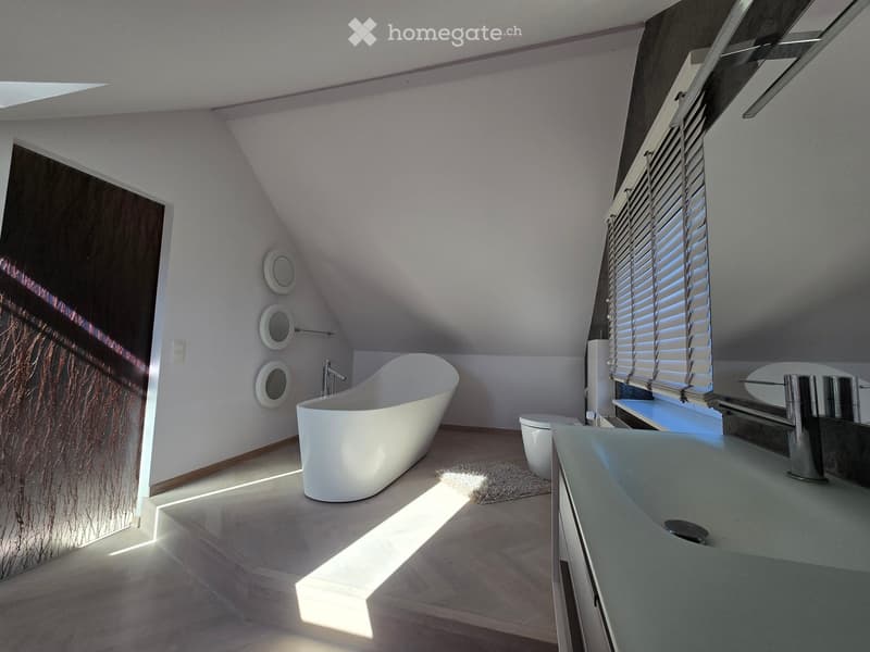 Modern bathroom with a freestanding bathtub, toilet, sink, and sleek blinds on the window. The room features light-colored walls and wooden flooring.