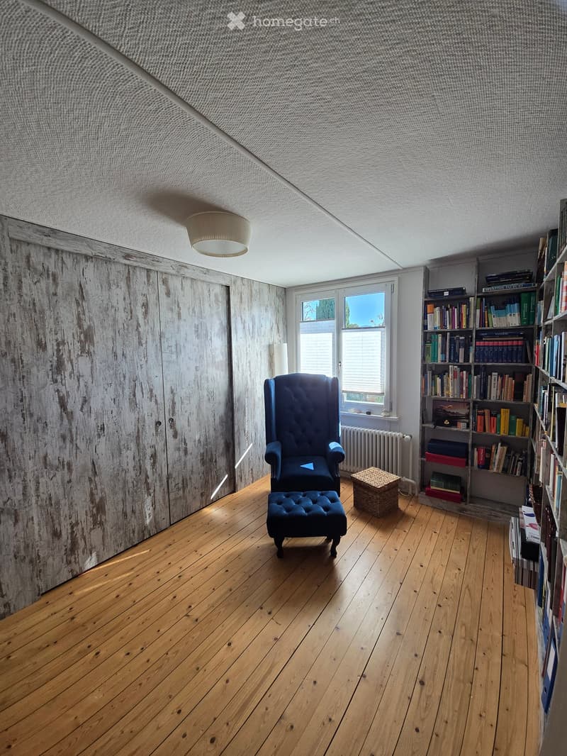 Cozy room with wooden floors, exposed wooden walls, large window, armchair, ottoman, bookshelf, and a woven basket.