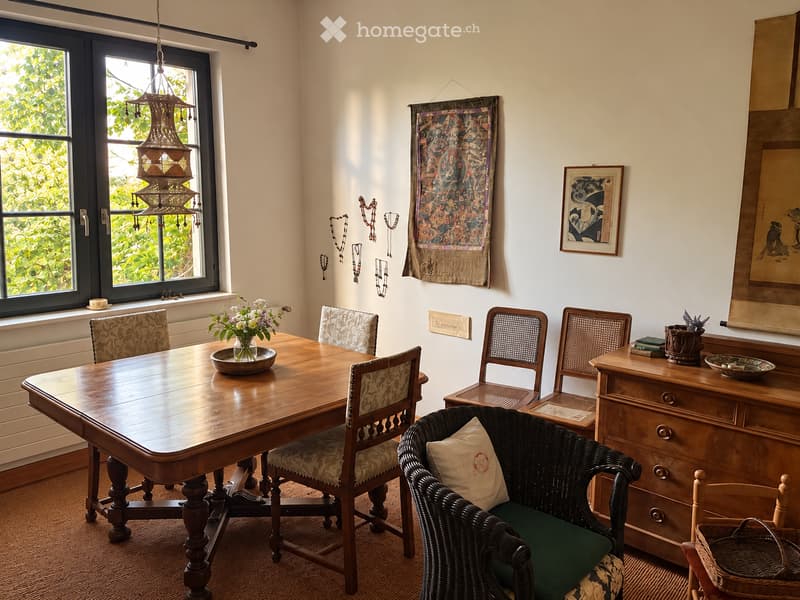 The image shows a well-furnished dining area with a wooden dining table surrounded by chairs, a wicker chair with a cushion, a vase with flowers, and decorative items on a side table.