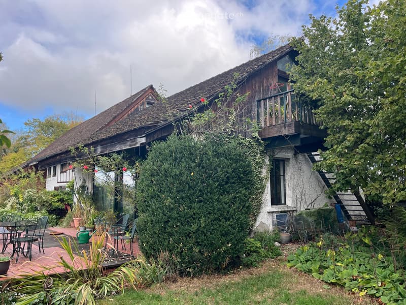 A rustic wooden house with a balcony, surrounded by lush greenery and a patio area with outdoor furniture.