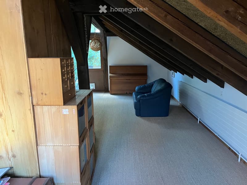 Attic room with exposed wooden beams, carpeted floor, and a slanted ceiling. Features a blue sofa, a wooden piano, a tall wooden cabinet, and a wicker pendant light.