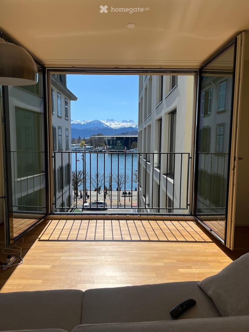 Bright living room with wooden flooring, large open sliding doors leading to a balcony with a scenic view of a lake and mountains. A grey sofa with a remote control is visible.