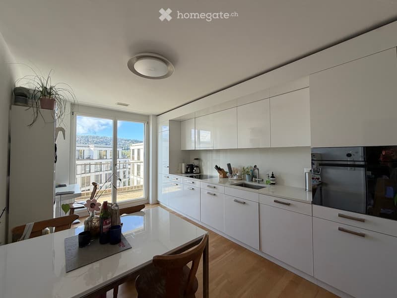 Modern kitchen with white cabinets, a central island, and a dining area. Features include a sink, oven, and a view of the balcony and cityscape.