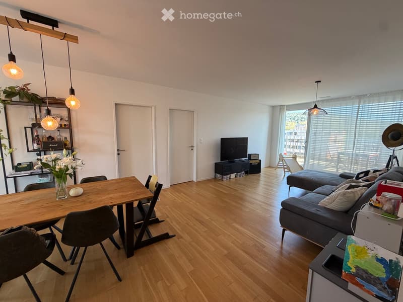 Modern living room with wooden flooring, a grey sectional sofa, a wooden dining table with chairs, a vase with white flowers, a television, and a shelf with decorative items.