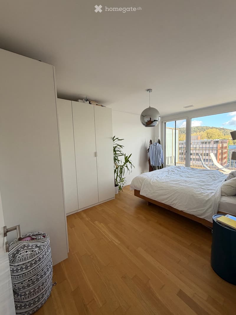 Modern bedroom with wooden flooring, large bed, wardrobe, potted plant, and a view of the landscape through large windows.