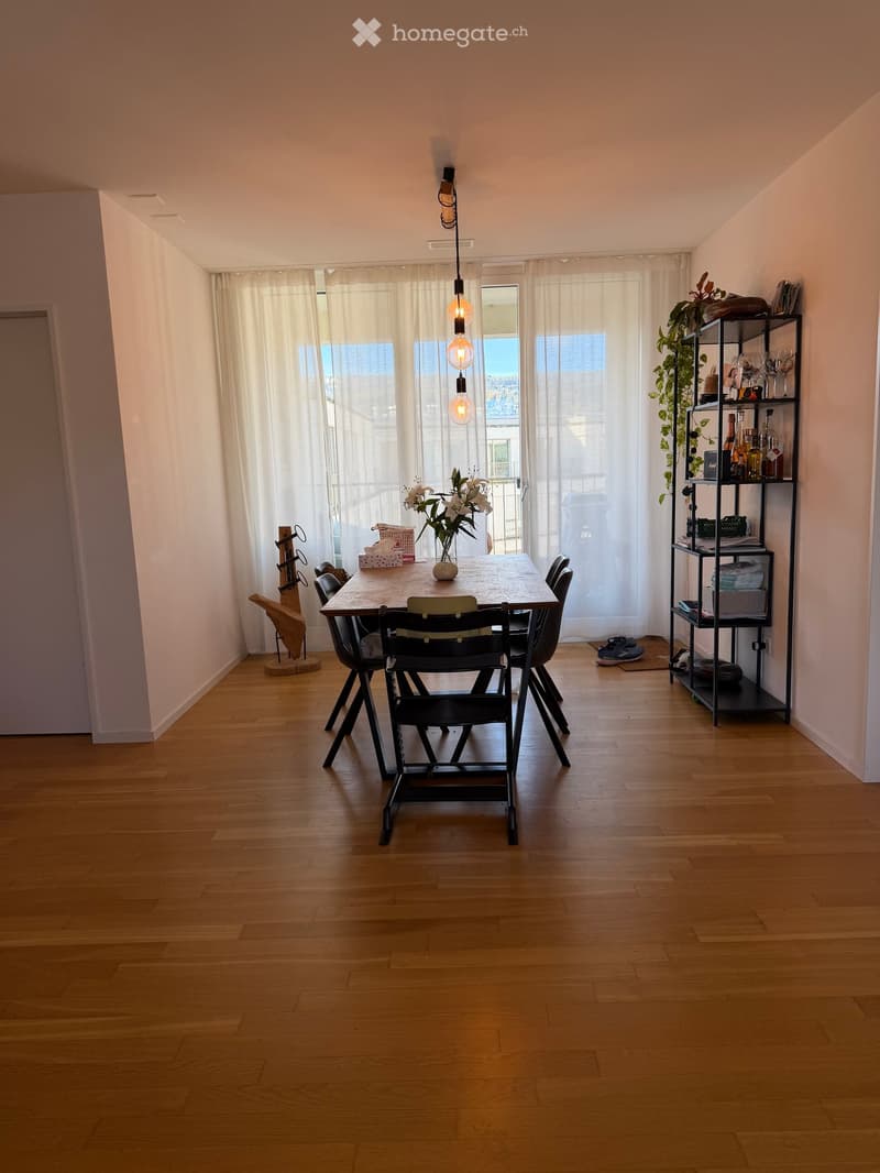 Dining area with wooden floor, large window with white curtains, wooden dining table with chairs, shelf with bottles and items, vase with flowers, and a hanging light fixture.