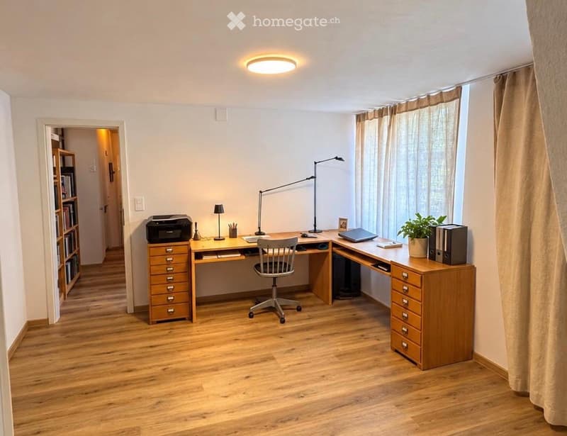A well-lit, modern home office with a large wooden desk, ergonomic chair, desk lamp, printer, laptop, potted plant, and filing cabinet. The room features light-colored walls, wooden flooring, and large windows with curtains.