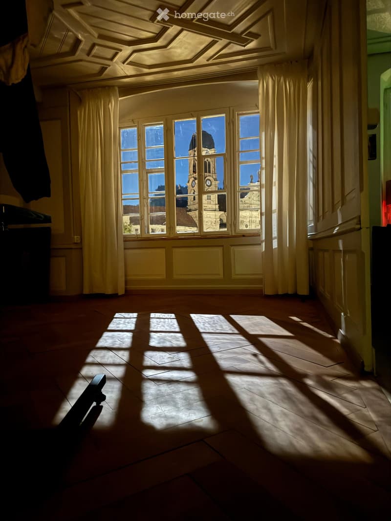 Empty room with wooden floor, large windows with curtains, and ornate ceiling. Sunlight streams in, casting shadows on the floor.