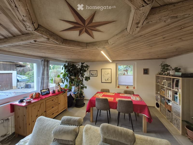 Cozy living and dining area with exposed wooden beams, a large star-shaped light fixture, a sofa, a dining table with chairs, a cabinet, a bookshelf, and a large potted plant.