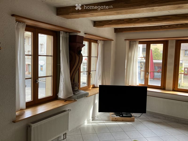 Bright living room with large windows, wooden beams on the ceiling, tiled floor, television on a wooden stand, and a small table with a decorative item on a window sill.