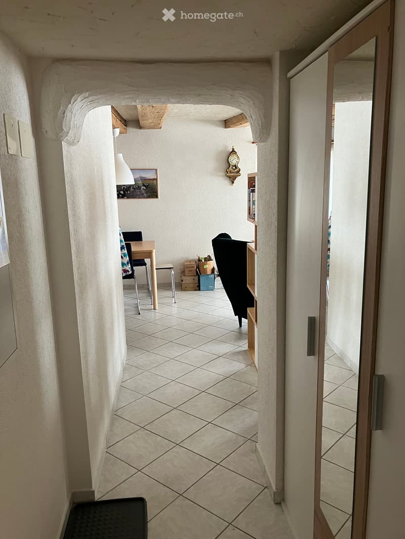 A narrow hallway with tiled flooring, white walls, and a wooden door leading to another room. Visible objects include a small table, chairs, a lamp, and a clock on the wall.