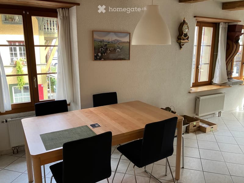 Dining area with a wooden table, four black chairs, a picture frame on the wall, a clock, and radiators under the windows.