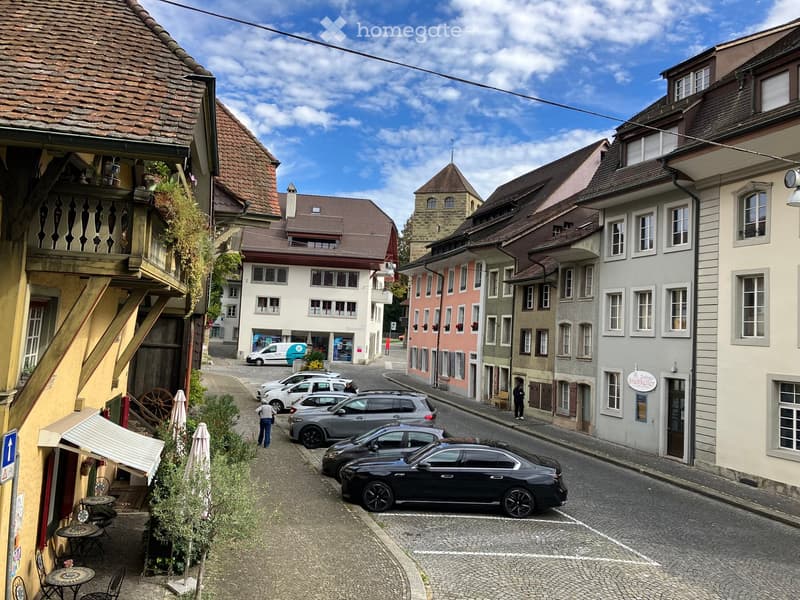 A picturesque European town street lined with colorful buildings, featuring a mix of architectural styles, parked cars, and a few pedestrians.