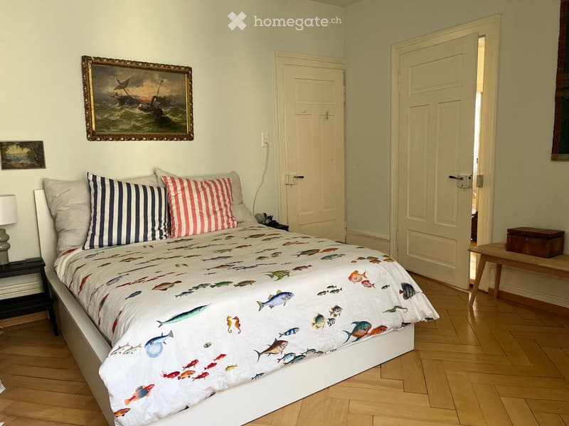 Cozy bedroom with wooden flooring, a large bed with a fish-patterned duvet, nightstand with lamp, and a painting above the bed.