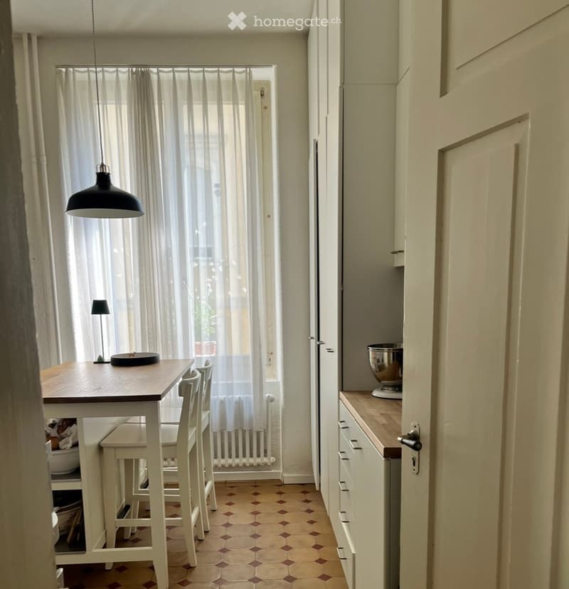 Bright kitchen with white cabinets, wooden table, and four chairs. A hanging lamp and a small table lamp are visible, along with a bowl on the counter and a bowl on the table.