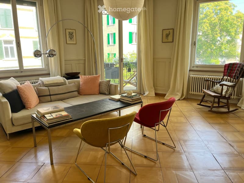 Living room with a light-colored sofa, coffee table, chairs, books, and a large window with curtains.