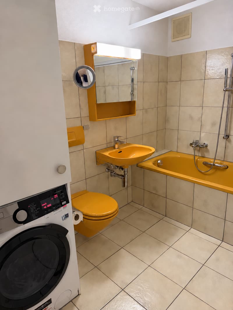 Modern bathroom with beige tiles, yellow fixtures including a sink, toilet, and bathtub, a mirror with a built-in shelf, and a washing machine.