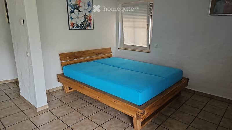 A simple bedroom with a wooden bed frame and blue mattress, tiled floor, and a window with blinds.