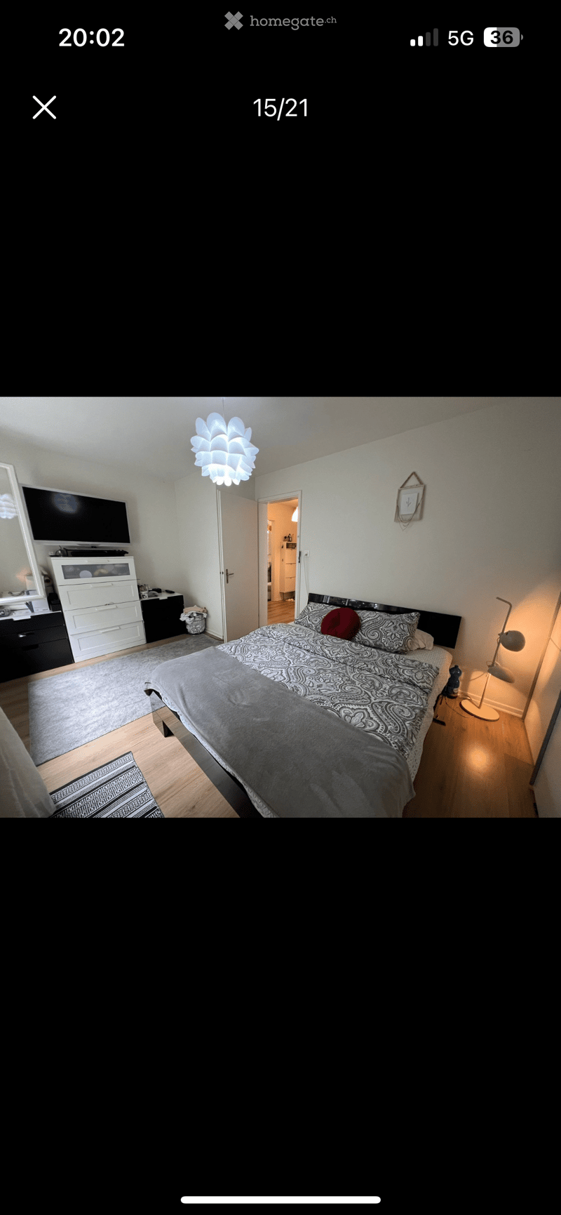 Well-lit bedroom with a large bed, patterned bedding, nightstand with lamp, dresser, TV, and a mirror on the wall.