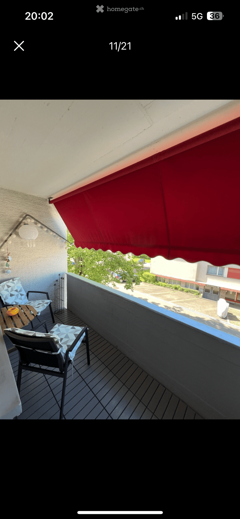 Balcony with wooden decking, black railing, and a red awning. Includes a small table and two chairs with geometric-patterned cushions.