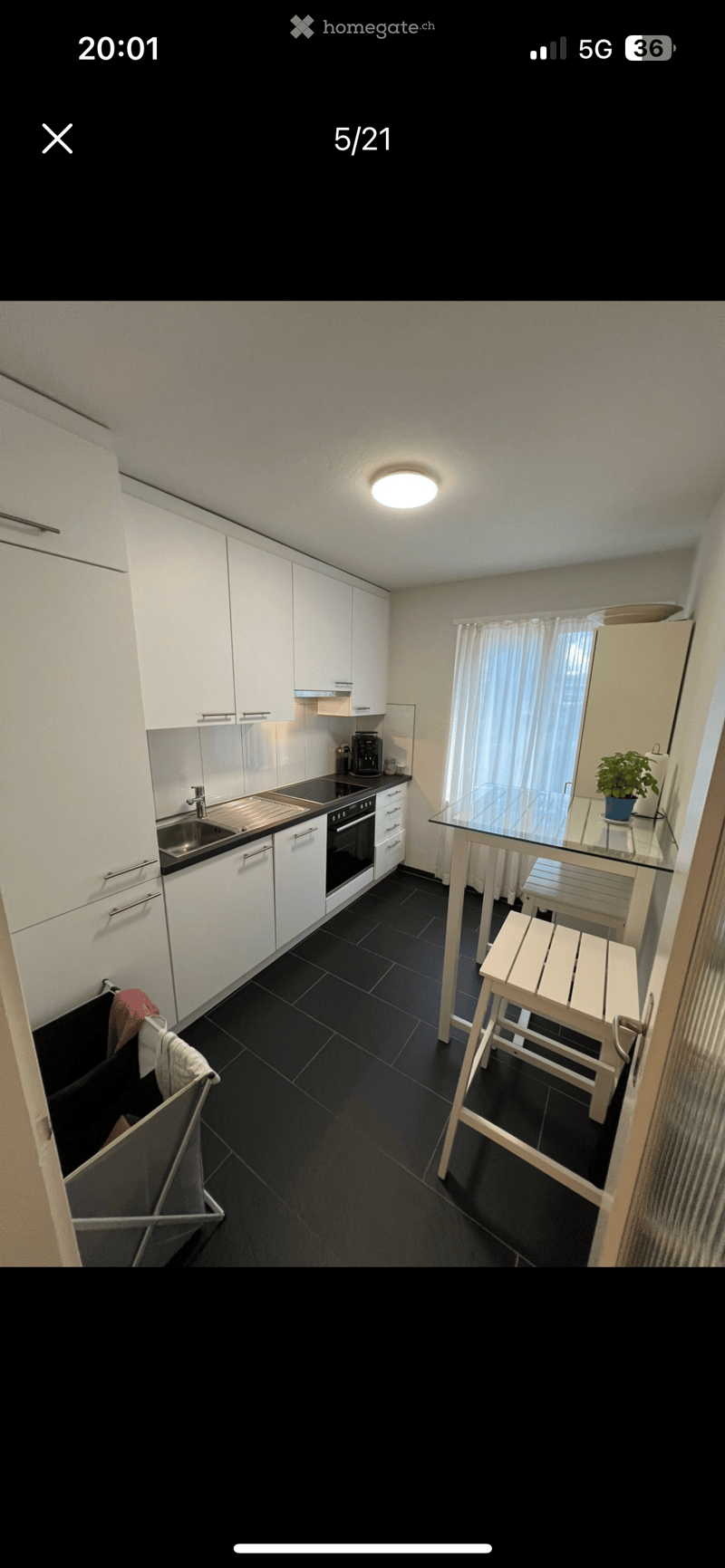 Modern kitchen with white cabinets, black tiled floor, stainless steel sink, oven, coffee maker, and a small dining area with a glass table and wooden stools.