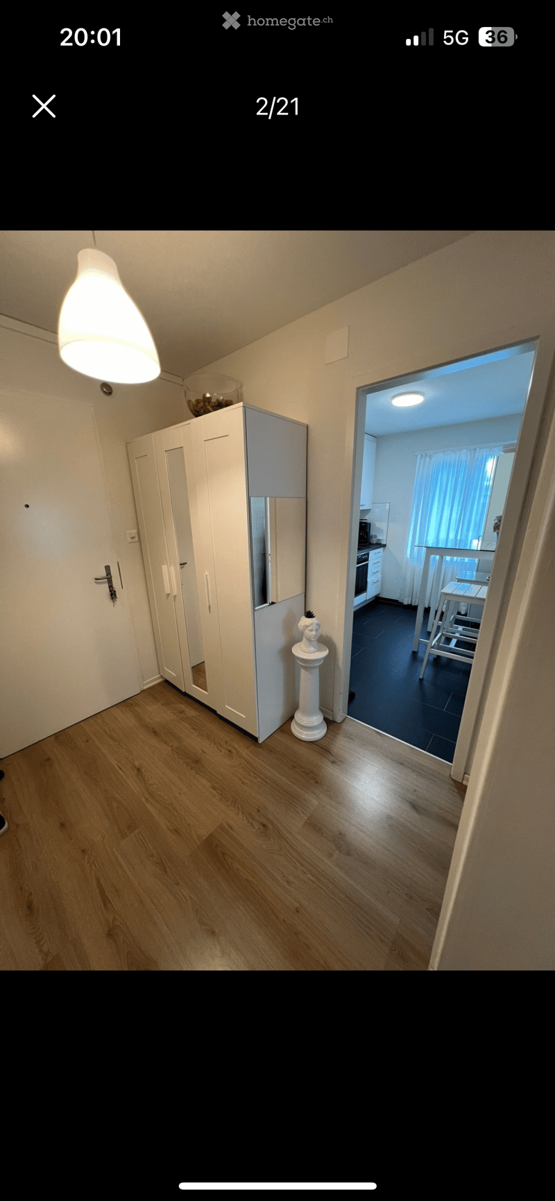 A well-lit hallway with wooden flooring, a large white wardrobe with mirrored doors, a decorative pedestal with a bust, and an open doorway leading to a modern kitchen area with white cabinets, a countertop, and bar stools.