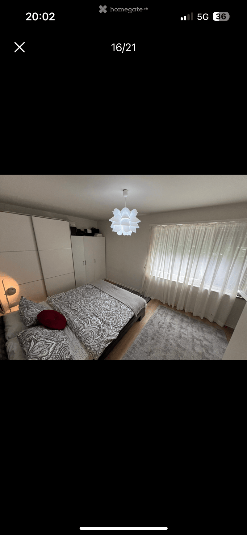 A well-lit bedroom with a bed featuring patterned bedding and pillows, a wardrobe, a large window with curtains, a rug, and a modern hanging light fixture.