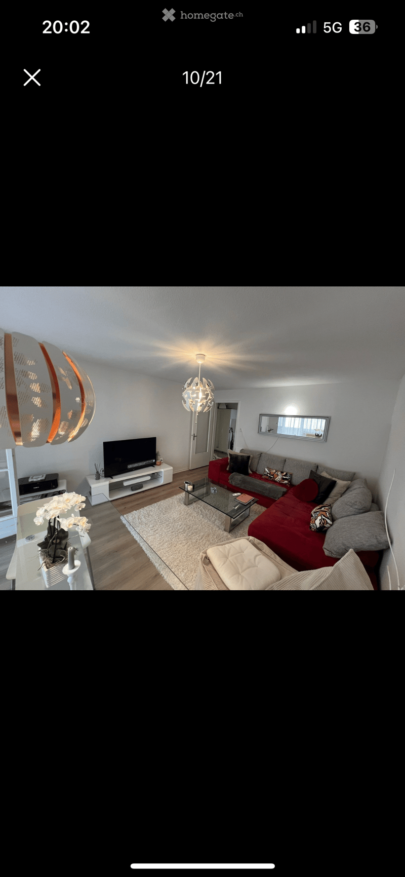 Modern living room with a red sectional sofa, glass coffee table, ottoman, flat-screen TV on a white stand, and decorative flower vase on a side table. The room features a white shag rug, wooden flooring, and a stylish chandelier.