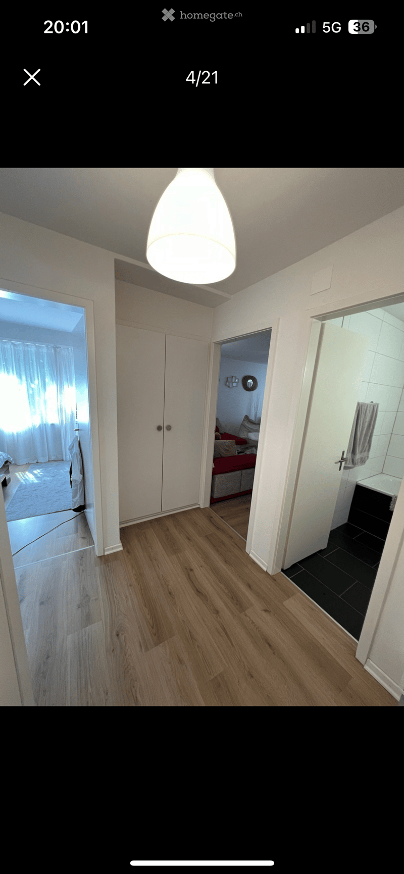 Empty hallway with wooden flooring, white walls, and a modern pendant light fixture. Doors lead to a bedroom and a bathroom.