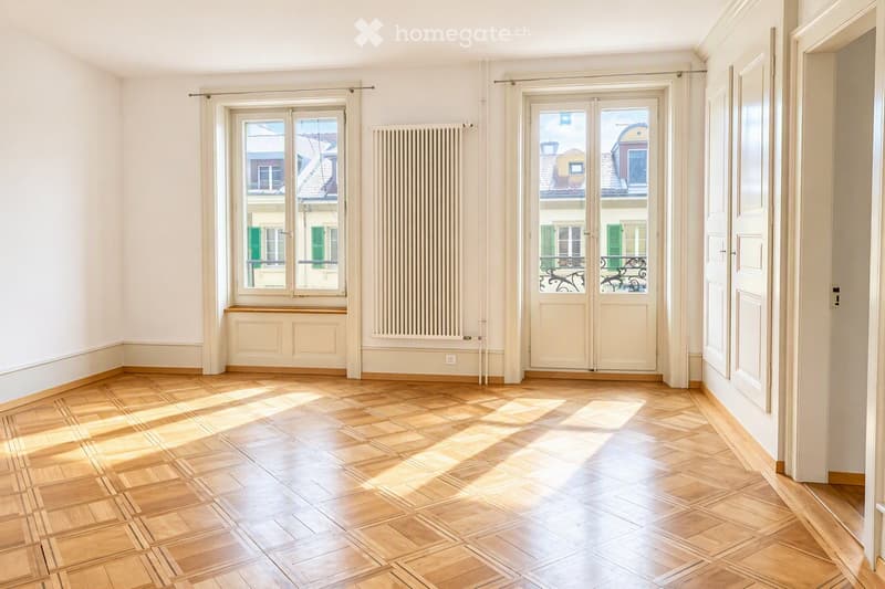 Empty room with wooden parquet flooring, white walls, large windows, and a radiator. No furniture or equipment present.