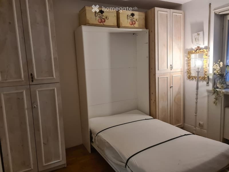 A small bedroom with a single bed, white wardrobe, mirror, and a decorative lamp near the window.