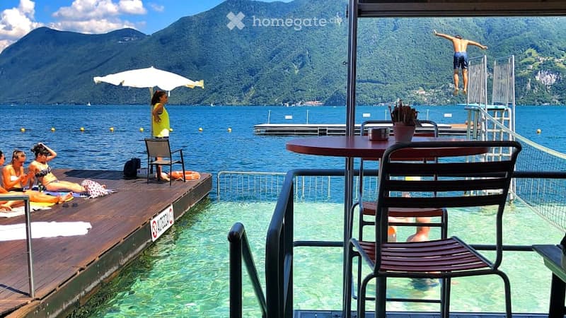 Outdoor area by a lake with wooden deck, lounge chairs, umbrella, and a diving board with a person jumping into the water. Scenic mountain view in the background.
