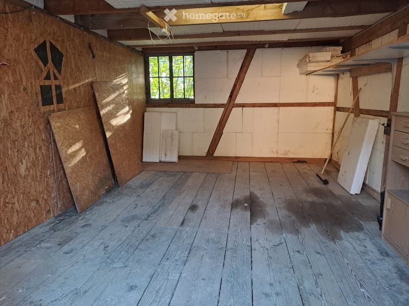 Empty room with wooden floors, exposed wooden beams, and a single window. Walls are unfinished with visible insulation and wooden panels. No furniture or equipment is present.