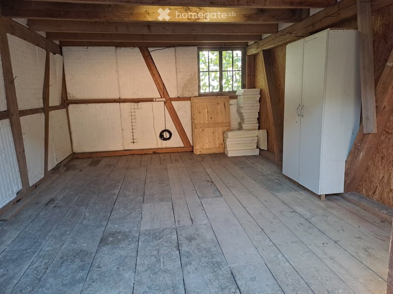 Empty room with wooden floor, exposed wooden beams, and a single window. There is a white cabinet and some stacked materials in the corner.