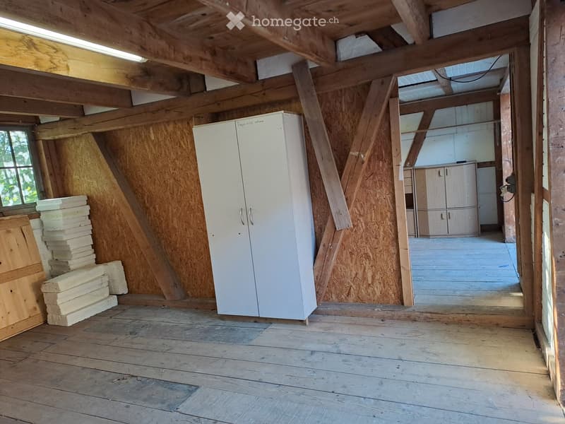 Empty room with wooden beams and flooring, featuring a white cabinet, stacked wooden planks, and a small window letting in natural light.